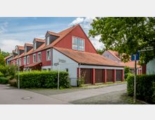Straßenansicht eines Wohnhauses mit roten Fassaden und Satteldächern aus roten Ziegeln, daneben eine angrenzende Garage mit mehreren braunen Toren. Im Vordergrund Gehwege und gepflegte Büsche, im Hintergrund Bäume und blauer Himmel.