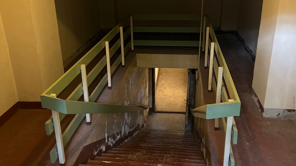 Blick in einen Innenraum-Treppenabgang mit breiter, abwärts führender Treppe, grün-weißen Geländern und abgenutztem rotbraunem Boden. Die Wände und Decke sind schlicht gestrichen, und unten führt ein schmaler Durchgang in einen weiteren Flur.