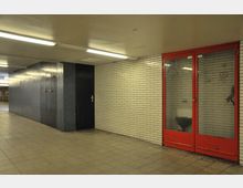 Unterirdischer Gang mit weißen Fliesenwänden, einer schwarzen Tür und einer Glasfront mit rotem Rahmen, hinter der sich ein Toilettenbereich befindet. Die Beleuchtung erfolgt durch längliche Deckenleuchten, und der Boden ist mit rechteckigen Fliesen ausgelegt.