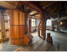 Innenraum einer historischen Mühle mit Holzbalkenkonstruktion. Im Vordergrund befindet sich ein großes, zylindrisches Holzsilo, während im Hintergrund Fenster mit natürlichem Licht und Ausstellungstafeln zu sehen sind.