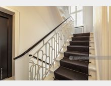 Treppenhaus in einem Gebäude mit weiß gestrichenem Geländer im dekorativen Bogenstil, dunklem Handlauf und einem Teppichläufer auf den Stufen. Im Hintergrund große Fenster, die Tageslicht hereinlassen.