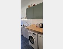 Kleine Küche mit einer Arbeitsplatte aus Holz, grauen Oberschränken und weißer Kachelrückwand. Unter der Arbeitsplatte befinden sich eine Waschmaschine und ein kleiner Kühlschrank; daneben stehen Spüle und Herd.