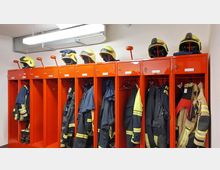 Rote Feuerwehrspinde mit Ausrüstungen, darunter Schutzanzüge, Stiefel und Helme mit gelben Abdeckungen. Auf den Spinden sind Beschriftungen angebracht, und die Umgebung ist ein heller, funktionaler Raum mit weißer Decke und sichtbaren Lüftungsrohren.