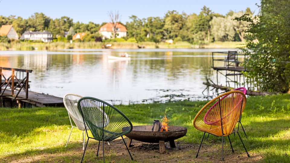 Uferwiese an einem ruhigen See mit drei farbigen Lounge-Stühlen um eine flache Feuerschale. Im Hintergrund sind Stege am Wasser sowie Häuser und Bäume am gegenüberliegenden Ufer zu sehen.