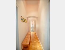 Langer, schmaler Flur mit hellem Holzboden und weißen Wänden. Links steht eine Holzbänksitzgruppe, und am Ende des Flurs ist ein Badezimmer mit weißen Fliesen sichtbar.