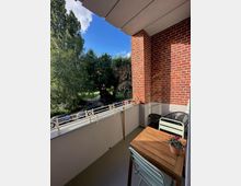 Ein Balkon mit einer roten Backsteinwand und weißem Geländer mit dekorativen Elementen. Im Vordergrund steht ein kleiner Holztisch mit zwei Stühlen und einer Topfpflanze, im Hintergrund ein Blick auf Bäume, parkende Autos und blauen Himmel.