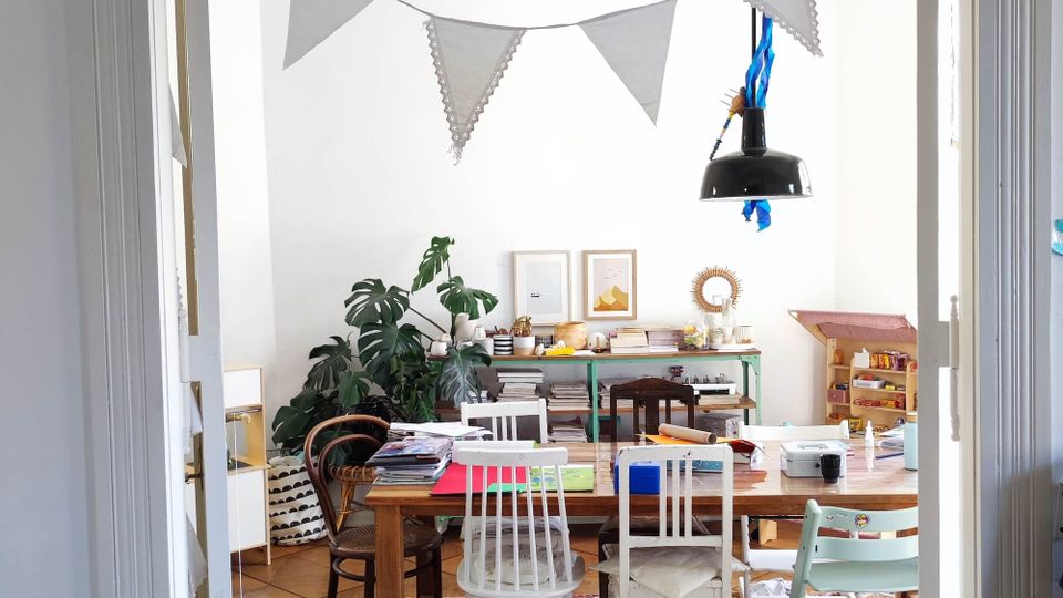 A cozy, cluttered workspace with a wooden table surrounded by mismatched chairs, shelves filled with books and art supplies, and green plants adding a touch of nature. A white wall features artwork, while a string of decorative pennants hangs from above, and a black pendant light shines over the table.