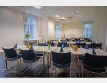 Heller Veranstaltungsraum mit mehreren gedeckten Tischen, blau gefalteten Stoffservietten und Gläsern. Große Fenster mit vertikalen Lamellen lassen Tageslicht herein, während der Raum mit weißen Wänden und dunklem Holzfußboden schlicht gestaltet ist.