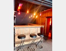 Eine minimalistisch gestaltete Bar mit hölzerner Verkleidung, drei schwarzen Barhockern mit Metallgestellen und freihängenden Glühbirnen, die warmes Licht abstrahlen. Im Hintergrund sind schwarze Wände und eine rote Neonleuchte sichtbar.