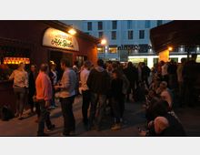 Eine Gruppe von Menschen versammelt sich vor einer roten Fassade mit einem Schild, auf dem "Bierstube Alt Berlin" steht, beleuchtet von warmem Licht. Im Hintergrund ist ein modernes Gebäude mit weißen Fassaden und großen Fenstern sichtbar.