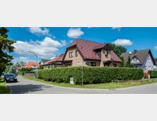 Wohnstraße mit Einfamilienhäusern, umgeben von gepflegten Hecken und Gärten. Im Vordergrund ein Ziegelhaus mit rotem Satteldach, links ein parkendes Auto und eine Straßenlaterne unter blauem Himmel mit Wolken.