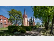 Backsteinkirche im neugotischen Stil mit gelber Fassade und schlankem Turm, umgeben von üppigem Grün und einer Parklandschaft; links ein rotes Backsteingebäude, rechts ein Weg, gesäumt von Bäumen.