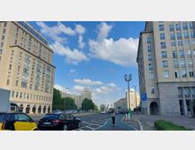 Straßenszene mit mehrstöckigen Gebäuden im Stil der Nachkriegsarchitektur auf beiden Seiten, flankiert von einer Fahrbahn und einem Radweg. Im Hintergrund sind Bäume und weitere Gebäudefassaden unter blauem, leicht bewölktem Himmel zu sehen.