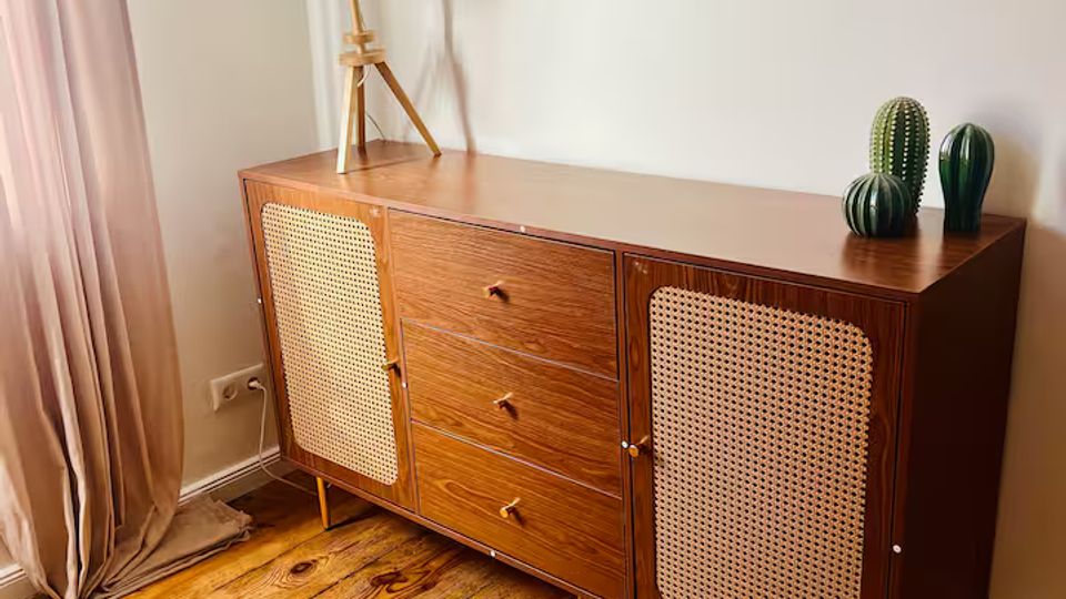 Heller Innenraum mit weißer Wand und breiten Holzdielen; davor steht eine niedrige Holzkommode mit zwei Rattantüren und drei Schubladen. Auf der Kommode befinden sich eine Dreibein-Tischlampe und zwei kleine Kaktus-Dekorationen, links hängt ein bodenlanger Vorhang.