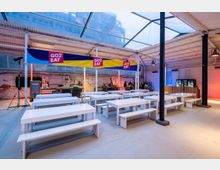 Ein heller, überdachter Gastrobereich mit weißen Holzbänken und Tischen, einer Bar im Hintergrund und bunten Bannern mit der Aufschrift "GO2EAT". Die Umgebung ist industriell gestaltet mit freiliegenden Backsteinwänden und einer Glasdachkonstruktion.