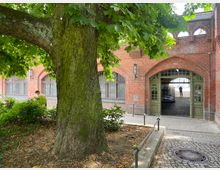 Großer Baum im Vordergrund mit dichtem Laub, daneben ein roter Backsteinbau mit Rundbogenfenstern und einem offenen Torbogen mit grünen Doppeltüren. Im Hintergrund ein gepflasterter Hof und vereinzelte Personen erkennbar.