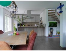 Heller, moderner Wohnraum mit offener Küche, Esstisch aus Holz und bunten Stühlen. Im Hintergrund eine Treppe, ein großes Aquarium und Kinderspielzeug; dekorative Hängelampen hängen über dem Tisch.