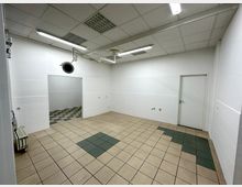 Ein leerer Raum mit beige- und dunkelgrün gemusterten Fliesenboden, weißen gefliesten Wänden und einer abgehängten Decke mit integriertem Beleuchtungssystem. Ein Durchgang führt in einen angrenzenden Raum, eine weiße Tür befindet sich an der rechten Wand.