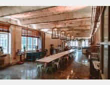 Industrieller Innenraum mit langen Holztischen und bunten Metallstühlen, umgeben von rohen Betonwänden, sichtbaren Rohrleitungen und großen Fenstern mit Holzjalousien. Der Raum ist mit modernen Käfiglampen beleuchtet und enthält dekorative Pflanzen sowie Arbeitsbereiche mit Vintage-Geräten.