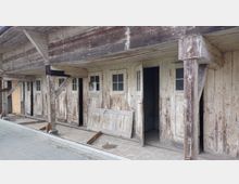 Holzgebäude mit verwitterten, weißen Türen und Fenstern, deren Glas teils beschädigt ist; das Gebäude zeigt deutliche Spuren von Verfall, mit abblätternder Farbe und sichtbaren Stützbalken aus Holz. Der Bereich vor dem Gebäude ist gepflastert, und eine beschädigte Holztür lehnt an der Fassade.