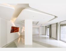 Moderner Innenraum mit weißen Wänden, geschwungener Decke und großen, deckenhohen Fenstern mit Vorhängen. Ein abgewinkeltes Fenster mit orangefarbenem Glas ist in die Wand integriert, der Boden ist glänzend und hell.