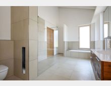 Modernes Badezimmer mit hellen Fliesen an Boden und Wänden. Zu sehen sind eine Toilette, eine Dusche mit Glaswand, eine Badewanne unter einem großen Fenster und ein Waschtisch mit Spiegel.