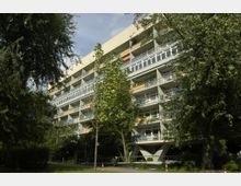 Ein mehrstöckiges Wohngebäude mit moderner Architektur, markant durch Balkone und große Fensterfronten. Das Gebäude ist von zahlreichen Bäumen umgeben, die in einer parkähnlichen Umgebung stehen.