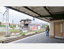 Bahnhof mit überdachtem Bahnsteig, Blick auf ein ehemaliges Stellwerk aus Ziegelstein, das mit Graffiti bedeckt ist. Im Hintergrund Gleise, Werbetafeln und moderne Gebäude; im Vordergrund zwei stehende Personen.
