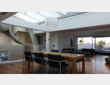 Helles, modernes Wohnzimmer mit Holzboden, einem rechteckigen Esstisch aus Holz und schwarzen Stühlen. Im Hintergrund gibt es eine geschwungene Treppe, eine große Fensterfront mit Zugang zur Terrasse und ein Glasdach, das viel Tageslicht hereinlässt.