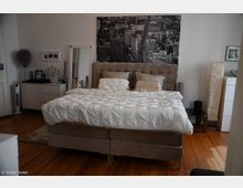 Ein Schlafzimmer mit einem großen Polsterbett, weißer Bettwäsche und mehreren Kissen. Im Hintergrund sind ein Schwarz-Weiß-Bild einer Stadtlandschaft, ein Spiegel, eine Kommode, sowie eine Stehlampe zu sehen; der Boden besteht aus hellem Holz.