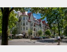 Straßenszene in einem Wohnviertel mit einem historischen Gebäudekomplex im Hintergrund, gekennzeichnet durch helle Fassade, rote Schrägdächer und zahlreiche Fenster. Umgeben von Bäumen, geparkten Autos und einer Ampel, fährt ein Radfahrer auf dem Radweg im Vordergrund.
