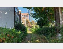 Zu sehen ist ein schmaler, bewachsener Weg zwischen überwucherten Sträuchern und Bäumen, flankiert von Gebäuden mit weiß verputzten Fassaden. Im Hintergrund sind moderne Bürogebäude mit Glas- und Backsteinfassaden sichtbar.