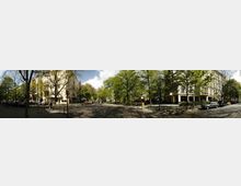 Straßenszene in einem städtischen Wohngebiet mit breiten Gehwegen und mehrstöckigen Wohnhäusern in heller Architektur. Große, dicht belaubte Bäume säumen die Straße, und geparkte Autos sowie vereinzelte Fußgänger sind sichtbar.