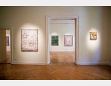 Ein Ausstellungsraum in einer Galerie mit hellen Wänden, Parkettboden und abstrakten Gemälden, die an den Wänden hängen. Durch eine offene Tür ist ein angrenzender Raum mit weiteren beleuchteten Gemälden sichtbar.