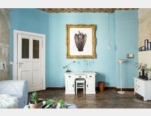 Wohnzimmer mit hellblauen Wänden, einer weißen Tür und einem Vintage-Schreibtisch mit Hocker. Ein goldgerahmtes Bild an der Wand, hölzerner Fischgrätboden und Dekorationen wie Zimmerpflanzen, Kerzenhalter und eine Stehlampe runden den Raum ab.