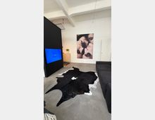 Modernes Loft-Wohnzimmer mit weißer Backsteinwand, Sichtbalkendecke und Betonboden. Ein schwarzes Trennelement mit Wandbildschirm, ein großes Wandbild, ein dunkles Sofa und ein schwarz-weißes Kuhfell als Teppich prägen den Raum.