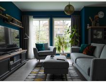 Wohnzimmer mit zwei grauen Sofas, einem passenden Hocker und einem niedrigen Fernsehtisch mit Flachbild-TV auf der linken Seite. Im Hintergrund befinden sich zwei große Fenster mit blickdichten Vorhängen, durch die Blick auf einen Garten und ein Haus gewährt wird; Zimmerpflanzen stehen vor den Fenstern. Die Wand ist dunkelblau gestrichen, und eine goldfarbene Deckenlampe hängt in der Mitte des Raumes. Ein farbiger Teppich mit geometrischen Mustern bedeckt den Boden.