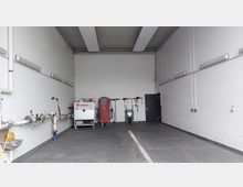 Ein leerer, rechteckiger Raum mit weißen, gefliesten Wänden, einer grauen Bodenfläche und energiesparenden Deckenleuchten. An der Rückwand befinden sich Maschinen, Reinigungsgeräte und Wandhaken mit Werkzeugen, während die linke Wand Wasserleitungen und ein Waschbecken zeigt.