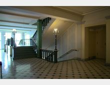 Eingangsbereich eines Gebäudes mit symmetrischem Fliesenboden aus achteckigen Mustern, einer zentralen Treppe mit schwarzem Geländer, grünen Säulen und Wandleuchten in Form von Kerzenständern. Tageslicht kommt durch große Fenster im Hintergrund.