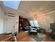 Wohnzimmer in einer Dachwohnung mit schrägen Wänden und Dachfenstern. Der Raum hat Holzboden, eine braune Eckcouch mit Teppich davor, eine moderne Deckenleuchte und eine Holzvertäfelung an einer Wand mit dekorativen Gegenständen.