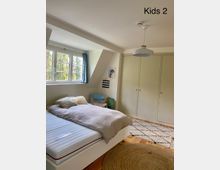 Kinderzimmer mit einem Bett links, großen weißen Einbauschränken rechts und einem Teppich mit geometrischem Muster auf Holzboden. Ein Fenster mit blauen Vorhängen lässt Tageslicht herein, und eine runde Hängeleuchte hängt an der Decke.