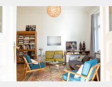Helles Wohnzimmer mit Holzboden, einem bunten Teppich und Retro-Möbeln wie blauen Sesseln, einer grauen Couch und einem gelben Zweisitzer. Im Hintergrund steht ein Bücherregal, an den Wänden hängen Bilder, und ein großer Kronleuchter aus Holz dominiert die Decke.