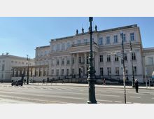 Historisches Gebäude mit klassizistischer Fassade, hohen Säulen und mehreren Fenstern in symmetrischer Anordnung an einer Straßenecke mit Straßenschildern und Laternen. Im Vordergrund ist der Fußweg und eine Fahrbahn zu sehen.