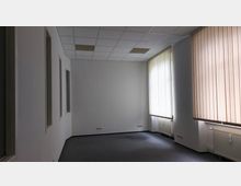 Leerer Büroraum mit weißen Wänden, grauem Teppichboden und einer abgehängten Decke mit eingebauten Leuchtpaneelen. Auf der rechten Seite befinden sich große Fenster mit vertikalen Jalousien, darunter Heizkörper.