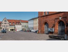 Historischer Marktplatz mit gepflastertem Boden, einer Reihe von mehrfarbigen Altstadthäusern mit Satteldächern im Hintergrund und einer großen roten Backsteinfassade mit Spitzbögen und Holzportalen im Vordergrund. Einige geparkte Autos und eine Statue in der Mitte des Platzes.