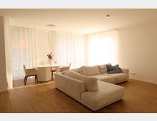 Helles Wohnzimmer mit Holzboden, einer großen beigefarbenen Eckcouch und einem runden Esstisch mit vier gepolsterten Stühlen. Große Fenster mit weißen, lichtdurchlässigen Vorhängen lassen viel Tageslicht herein, und eine moderne Hängelampe hängt über dem Tisch.