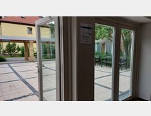 Blick durch eine Glastür auf einen gepflasterten Innenhof mit schwarzen Steinlinien, umrahmt von Pflanzen und Bänken; im Hintergrund ist ein gelbes Gebäude mit roten Dachziegeln zu sehen.