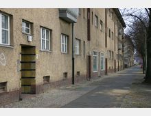 Mehrstöckiges Wohngebäude aus der Nachkriegszeit mit einer schlichten, beige gestrichenen Fassade und Backsteinelementen im unteren Bereich. Links führt ein schmaler Bürgersteig an der Häuserreihe entlang, flankiert von kahlen Bäumen.