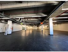 Weitläufiger Lager- oder Industriebereich mit unverputzten Betonwänden, freiliegenden Rohren und Lüftungsschächten an der Decke. Der Boden ist dunkel und glatt, gestützt von mehreren Betonpfeilern, und eine Leiter lehnt an einer der Wände.