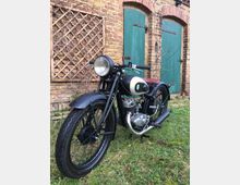 Ein schwarzes Oldtimer-Motorrad steht auf einer Rasenfläche vor einer Ziegelwand mit grünen Holztüren und Kletterpflanzen. Das Motorrad hat ein klassisches Design mit runden Scheinwerfern und einem Metallmotor.